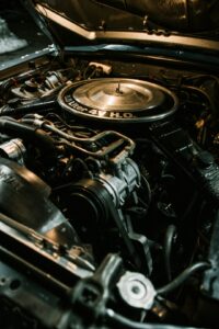 A mechanic examines the engine bay of a pickup truck next to a sedan in a residential driveway