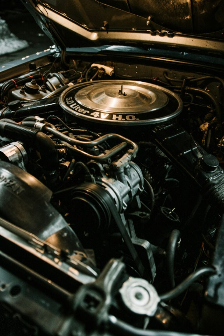 A mechanic examines the engine bay of a pickup truck next to a sedan in a residential driveway